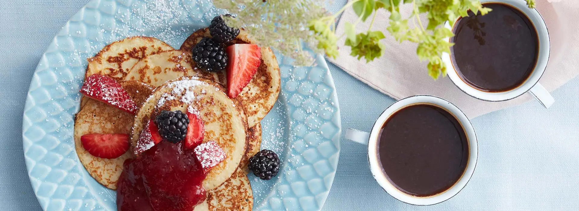Auf einem Teller liegen drei Pancakes mit Beeren, Puderzucker und Marmelade verziert. Daneben stehen zwei Tassen mit Kaffee.