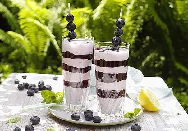 Zwei hohe Glasbecher zeigen einen gestreiften Blaubeershake mit abwechselnd lila und weißen Schichten, der bis zum Rand reicht. Auf jedem Glas balanciert ein Holzspieß mit frischen Blaubeeren, und an der Seite hängen bunte Strohhalme.