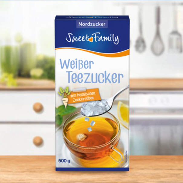 Eine Packung „Weißer Teezucker“ zeigt eine Tasse mit Tee und herabfallenden Zuckerkristallen; die Verpackung steht auf einer Küchenarbeitsplatte vor unscharfer Kulisse.