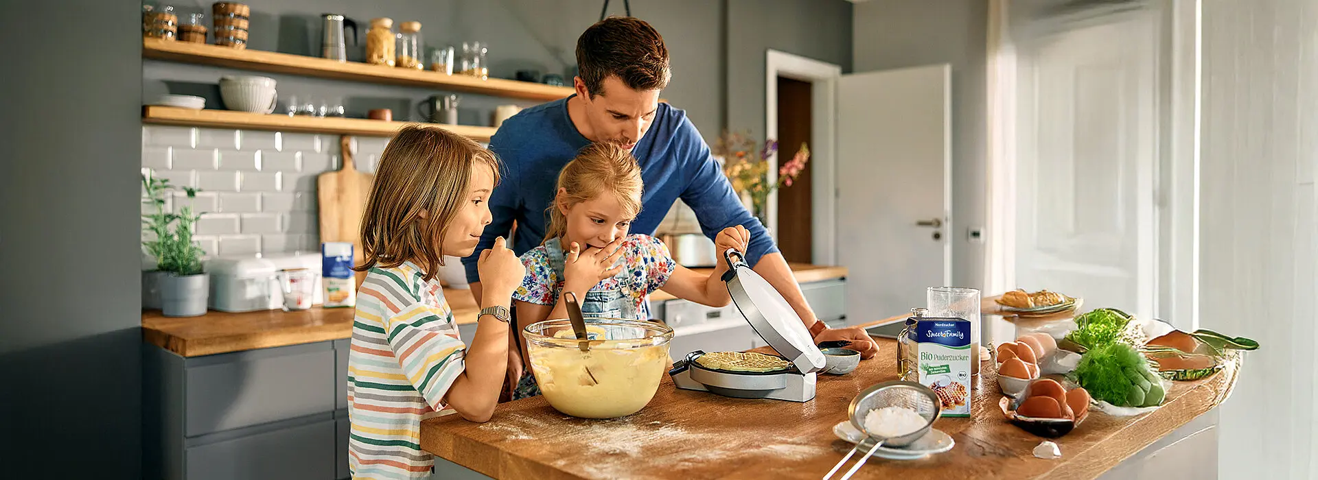 Ein Mann mit 2 Kindern backen in der Küche Waffeln und schauen gespannt auf das Ergebnis