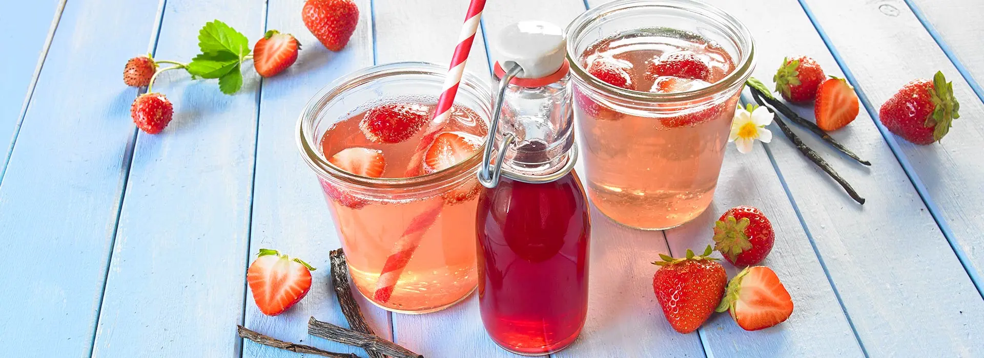 In zwei breiten Gläser, die ebenso gut Einmachgläser sein könntent, ist eine hell-rosane Limonade enthalten, in der Erdbeeren und ein rot gestreifter Strohhalm schwimmen. Davor steht eine kleine Glasflasche mit Sirup drin.
