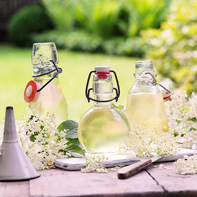 Auf einem alten Holztisch stehen mehrere Glasflaschen mit hellgelbem Holunderblütensirup, einige mit Bügelverschluss. Weiße Holunderblütendolden und grüne Blätter liegen locker neben den Flaschen, und ein kleiner Metalltrichter weist auf das Abfüllen hin.