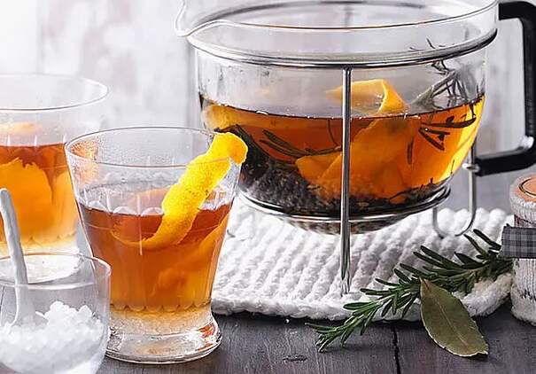 Eine gläserne Teekanne auf einem Stövchen ist mit einem dunklen Kräutertee gefüllt, in dem Orangen- und Zitronenschalen sowie Rosmarinzweige schwimmen.