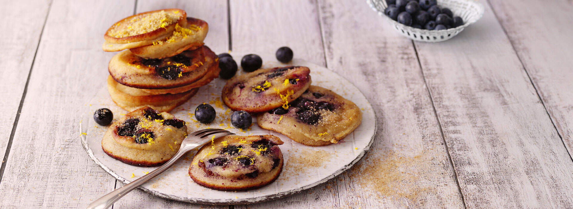 Auf einem weißen Porzellanteller sind mehrere kleine Kuchen arrangiert. Die Kuchen haben eine goldbraune Oberfläche und sind mit einer glänzenden, dunklen Blaubeerfüllung und gelber Zitronenglasurverzierung dekoriert. Ein Stapel von zwei Kuchen steht erhöht im Hintergrund, während die anderen Kuchen flach auf dem Teller liegen. Verstreute frische Blaubeeren und ein silberner Servierlöffel ergänzen die Szene. Im Hintergrund ist eine kleine Schale mit weiteren Blaubeeren zu sehen.