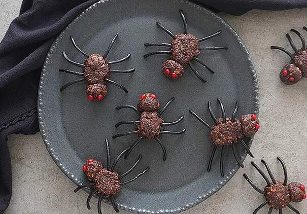 Auf einem dunklen, runden Tablett mit silbernem Rand sind Spinnen arrangiert. Jede Spinne hat einen glänzenden, braunen Körper aus Teig mit feinen, schwarzen Lakritzen als Beine. Einige Spinnen haben rote Zuckerstreusel als Akzente. Die Anordnung wirkt kunstvoll und ist perfekt für eine Halloween- oder Herbst-Dekoration.