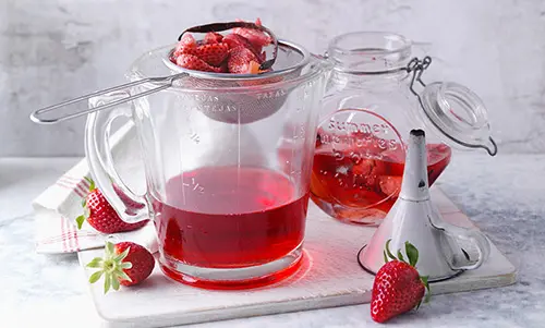 Auf einem großen Becher liegt ein feines Sieb auf dem eingemachte Erdbeeren liegen. 