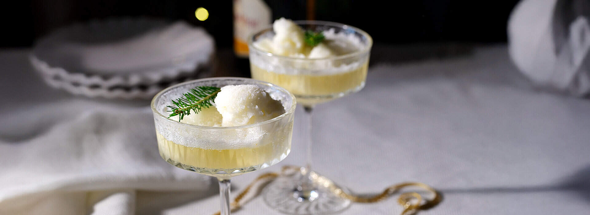 In einem flachen Coupeglas befindet sich eine helle Gelée mit einem Klecks Sorbet; ein grünes Zweigchen Thymian dient als Dekoration