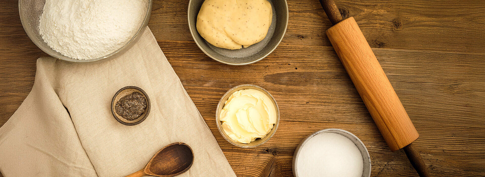Auf einer Holzplatte liegen Schalen mit Mehl, Zucker und veganer Margarine; ein Stück Teig und ein Nudelholz ergänzen die Szene, daneben befindet sich ein Holzlöffel sowie ein kleines Schälchen mit gemahlenen Samen.