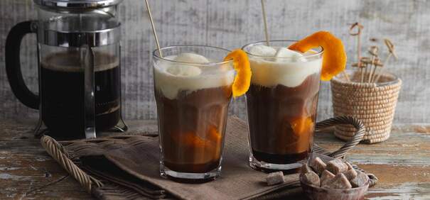 Zwei schlanke Gläser mit kaltem Kaffeegetränk sind mit Schaum bedeckt; je ein Orangenschnitz steckt am Glasrand
