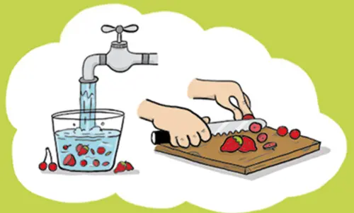 Aus einem Waaserhahn läuft Wasser in ein großes Glas mit Früchten drin.  Rechts daneben liegt ein Brett auf dem Erdbeeren geschnitten werden.