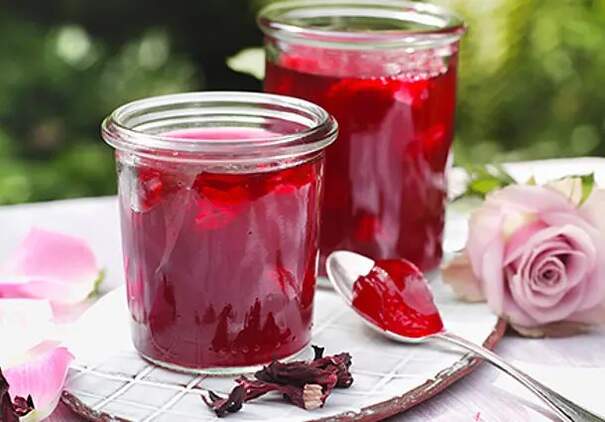 Ein kleines Glas mit leuchtend rotem Rosen‑Hibiskus‑Gelee steht auf einem hölzernen Untergrund, daneben steht ein weiteres Glas. Ein Silberlöffel hebt eine Portion des klaren Gelees heraus, sodass seine samtige Struktur sichtbar wird.