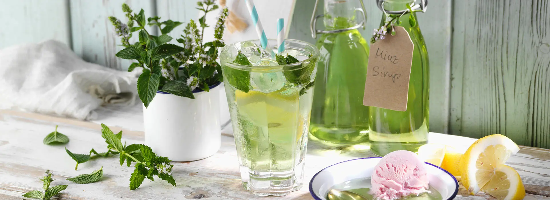 Auf einem Tisch stehen links zwei Glasflaschen, die eine grüne Flüssigkeit enthalten. Davor steht ein Glas, mit der gleichen Flüssigkeit. außerdem sind noch Pflanzen und eine Schale mit Löffel auf dem Tisch zu sehen. Das Szenario sieht nach Frühling aus.