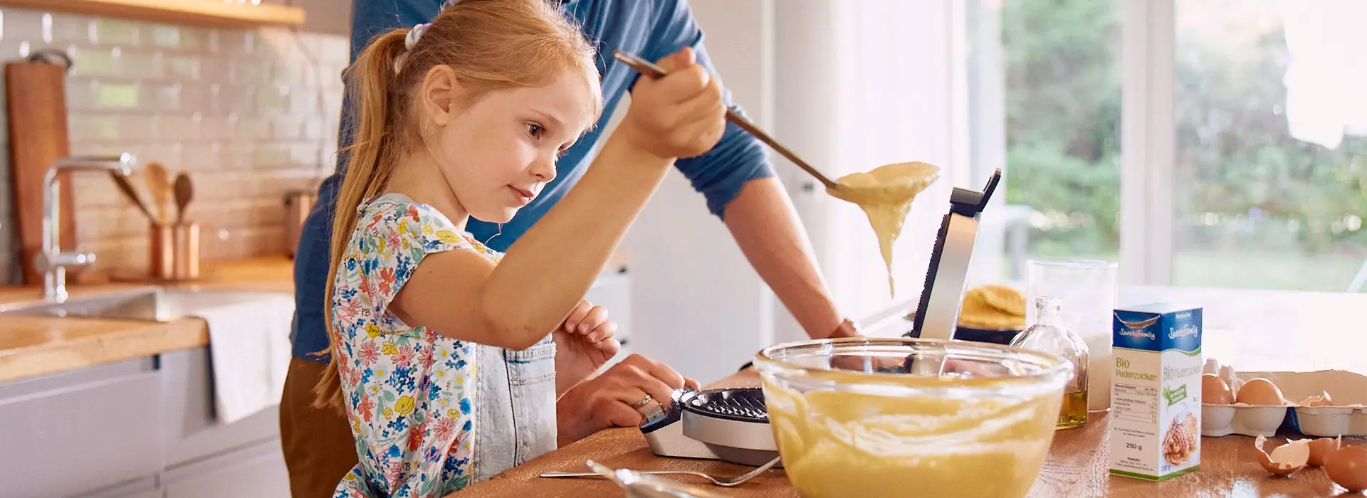 Mädchen steht in der Küche und schöpft Waffelteig mit einer großen Kelle von der Schüssel in ein Waffeleisen