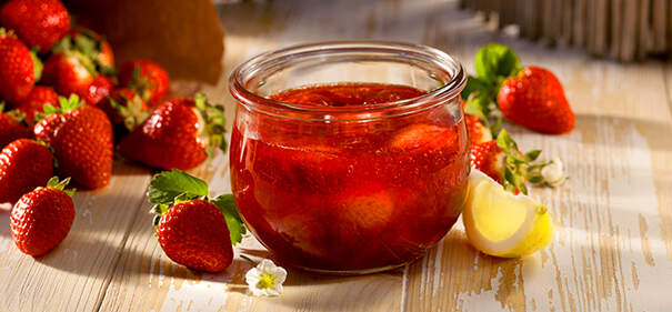 Ein Glas Erdbeermarmelade steht auf dem Tisch, daneben frische Erdbeeren und eine Zitronenspalte.
