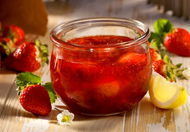 Rustikales Glas mit glänzender Erdbeermarmelade, daneben frische Erdbeeren und ein Löffel auf einem Holzbrett