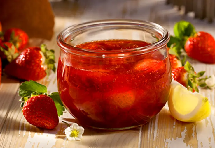 Rustikales Glas mit glänzender Erdbeermarmelade, daneben frische Erdbeeren und ein Löffel auf einem Holzbrett