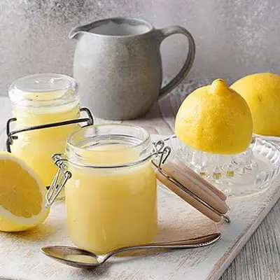 Zwei Weckgläser mit gelbem Fruchtaufstrich aus Zitronen. Daneben liegen frische Zitronen und eine Karaffe aus Ton steht im Hintergrund