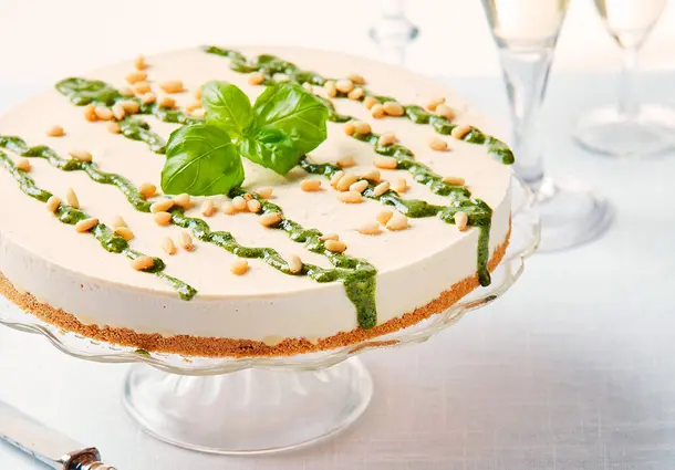 Ein gefrorener Zitronen‑Cheesecake steht auf einer Glasplatte und ist mit grünem Basilikum‑Pesto, Pinienkernen und einem Basilikumzweig garniert