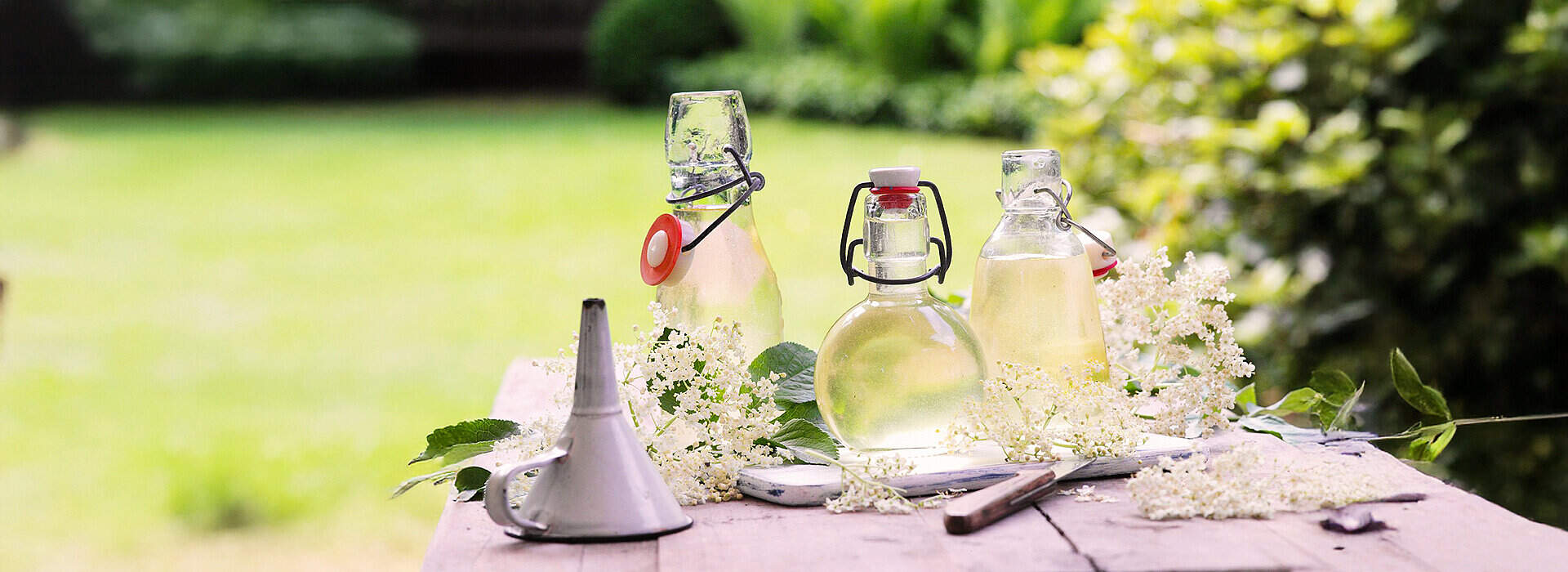 Auf einem alten Holztisch stehen mehrere Glasflaschen mit hellgelbem Holunderblütensirup, einige mit Bügelverschluss. Weiße Holunderblütendolden und grüne Blätter liegen locker neben den Flaschen, und ein kleiner Metalltrichter weist auf das Abfüllen hin.