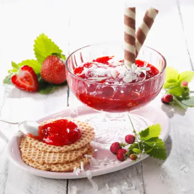In einem flachen Dessertglas ist eine rote Masse drin, verziert mit weißer Schokolade und Waffelrollen. Der Becher steht auf einem Teller auf dem 2 kleine Waffeln liegen.