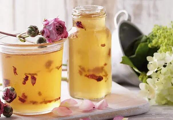 Zwei Gläser mit hellem Apfel‑Traubengelee sind liebevoll mit rosa Rosenblättern verziert. Daneben liegen kleine Rosenknospen und weiße Blüten, die dem Ensemble eine zarte Eleganz verleihen.
