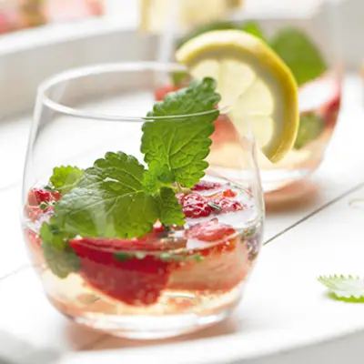 Ein heller Cocktail in einem bauchigen tiefen Glas, mit Himbeeren und Minze verziert. 