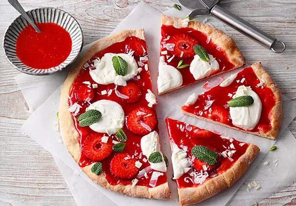 Auf einem hellen Holztisch liegt eine runde süße Pizza mit leuchtend rotem Erdbeerbelag, die in Stücke geschnitten ist. Jedes Stück ist mit weißen Sahnetupfen und grünen Minzblättern dekoriert, einige haben zusätzlich frische Erdbeerscheiben.