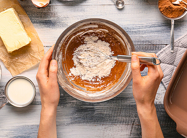 Blick von oben in eine Teigschüssel. Diese wird von zwei Händen gehalten und gerade wird Mehl unter eine Masse verrrüht. Daneben liegt ein Klotz Butter