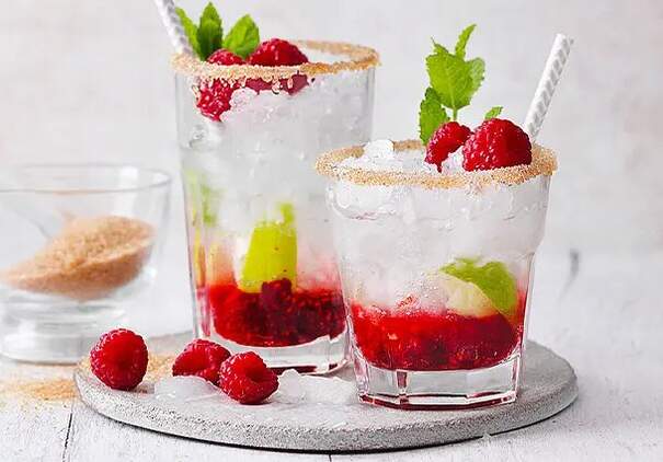 Zwei bauchige Cocktailgläser sind randvoll mit einer rosaroten Himbeer‑Caipirinha, die mit zerstoßenem Eis gefüllt ist. Die Glasränder sind mit Zucker bestreut; im Inneren schwimmen frische Himbeeren, Limettenscheiben und Minzblätter. 