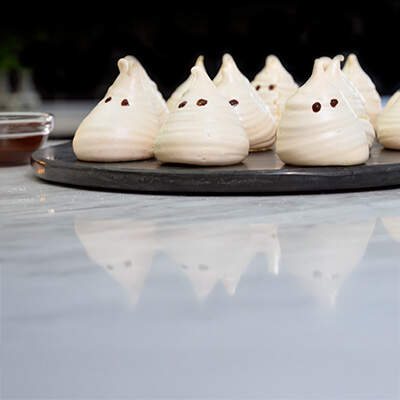 weiße Meringue-Geister in einer Reihe angeordnet. Jeder Geist hat eine charakteristische Spitzform mit zwei dunklen Schokoladenpunkten als Augen und einen dünnen, gewellten Schokoladenstrang als Mund. 