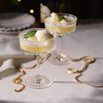 Auf dem Bild sieht man ein hohes, rundes Cocktailglas, das zur Hälfte mit einem gelben Cocktail gefüllt ist. Im Glas findet sich zudem eine Kugel Eis und ein kleiner Tannenzweig liegt dekorativ darüber. 