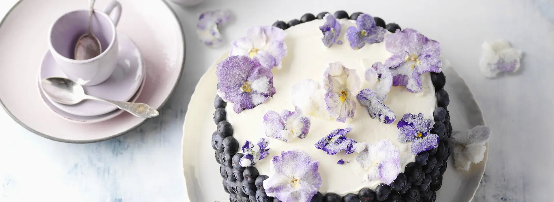 Ein lila Teeservice mit silbernen Teelöfflen steht neben einer hohen Cremetorte, die rundum mit Blaubeeren vestückt ist und oben drauf mit Zucker verzierte Veilchen in lila.
