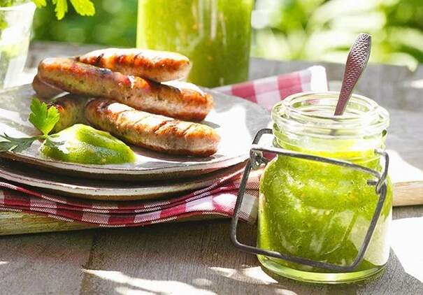 Ein Glas grüne Zucchini‑Senf‑Sauce steht neben gegrillten Würstchen und einer Limettenspalte.