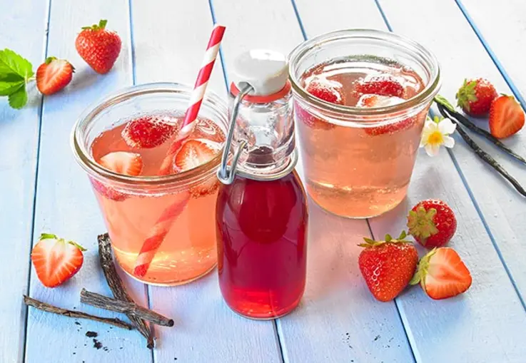 In zwei breiten Gläser, die ebenso gut Einmachgläser sein könntent, ist eine hell-rosane Limonade enthalten, in der Erdbeeren und ein rot gestreifter Strohhalm schwimmen. Davor steht eine kleine Glasflasche mit Sirup drin.