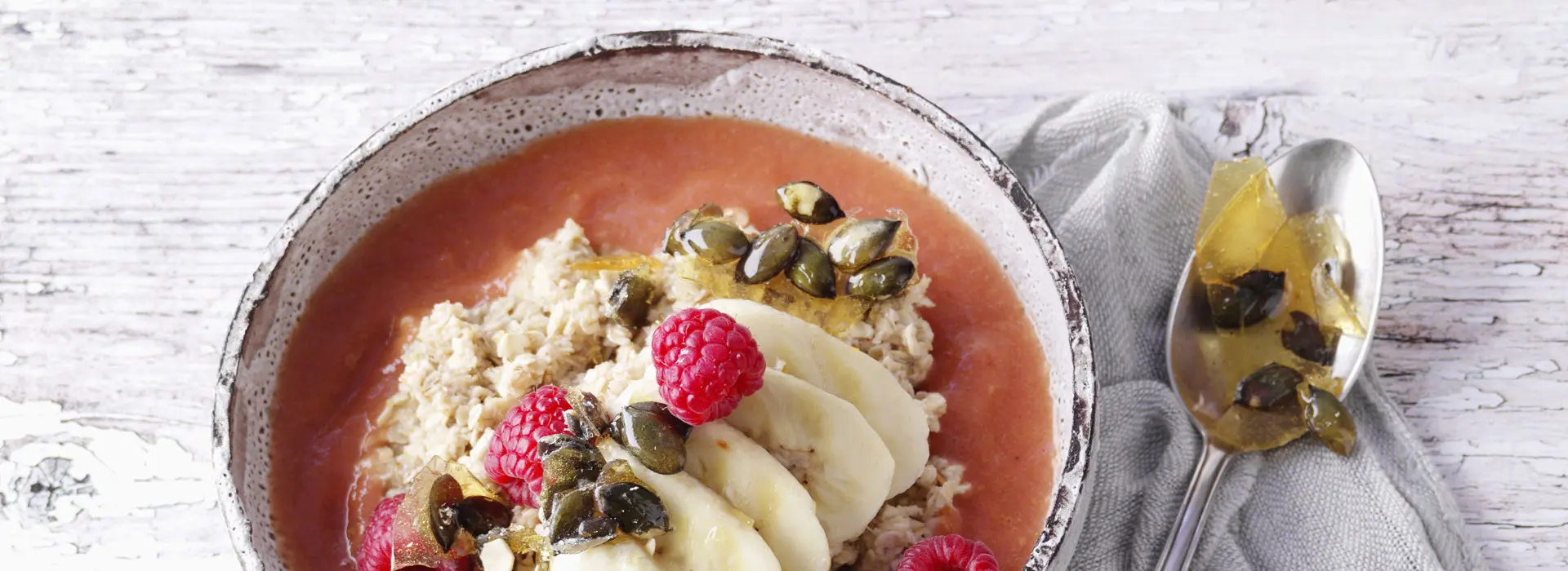 Eine Schale Porridge mit Bananenscheiben, Himbeeren, Kürbiskernen und Beerensoße.