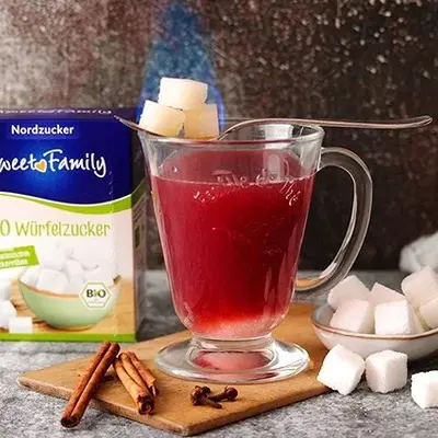 Eine heiße Tasse Punch, auf der ein Löffel liegt, der drei Würfelzucker trägt. Im Hintergrund die Verpackung des Würfelzuckers von Sweetfamily