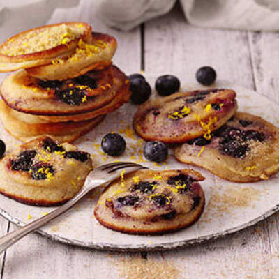 Auf einem weißen Porzellanteller sind mehrere kleine Kuchen arrangiert. Die Kuchen haben eine goldbraune Oberfläche und sind mit einer glänzenden, dunklen Blaubeerfüllung und gelber Zitronenglasurverzierung dekoriert. Ein Stapel von zwei Kuchen steht erhöht im Hintergrund, während die anderen Kuchen flach auf dem Teller liegen. Verstreute frische Blaubeeren und ein silberner Servierlöffel ergänzen die Szene. Im Hintergrund ist eine kleine Schale mit weiteren Blaubeeren zu sehen.