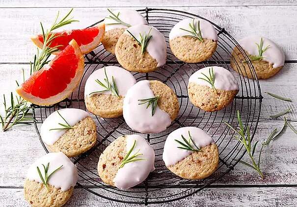 Runde Kekse auf einem Kuchenrost sind mit weißer Glasur und Rosmarinzweigen verziert. Auf dem Rand des Kuchenrosts liegen zudem zwei Viertel einer leuchtend roten Grapefruit.