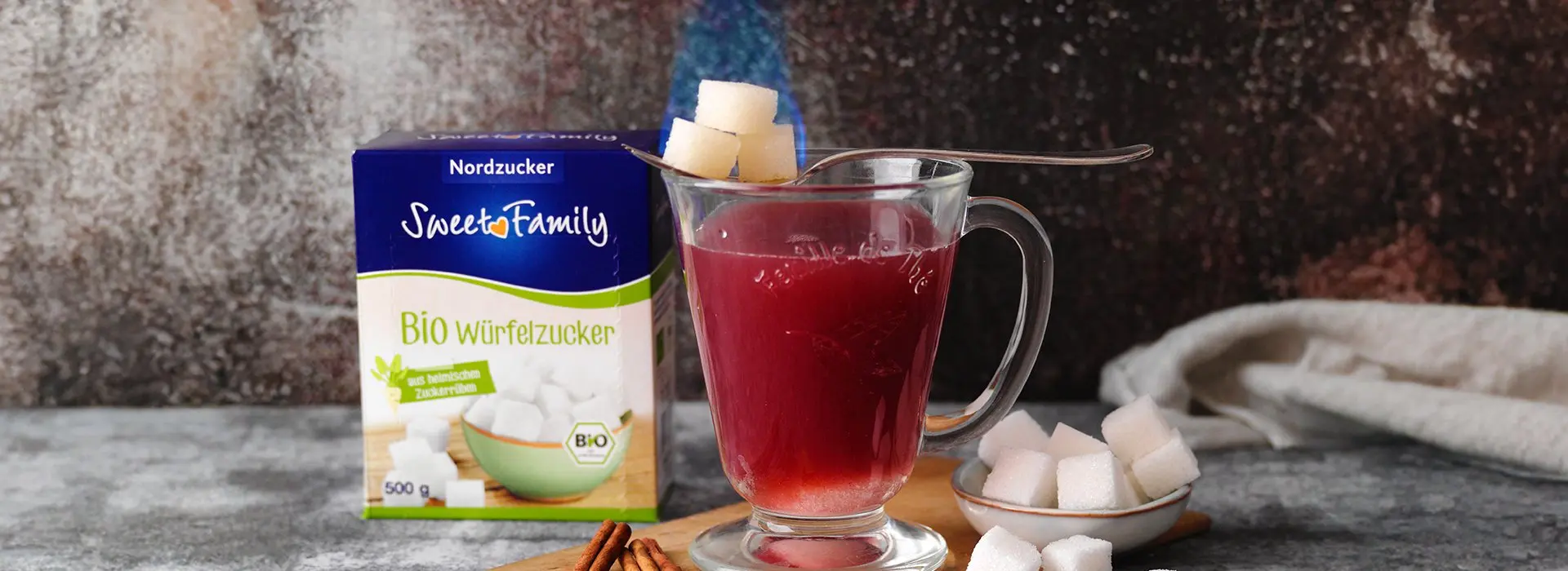 Eine heiße Tasse Punch, auf der ein Löffel liegt, der drei Würfelzucker trägt. Im Hintergrund die Verpackung des Würfelzuckers von Sweetfamily