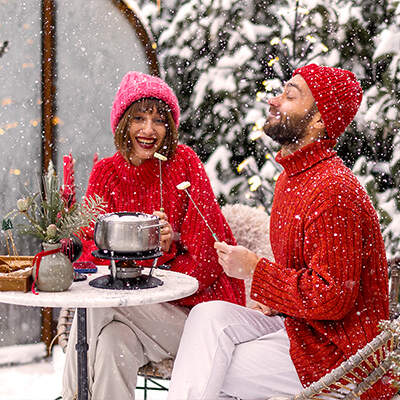 Ein Paar in roten Winterkleidern sitzt im verschneiten Garten; sie lächeln, während sie an einer Feuerschale oder einem kleinen Tisch mit dampfendem Getränk sitzen