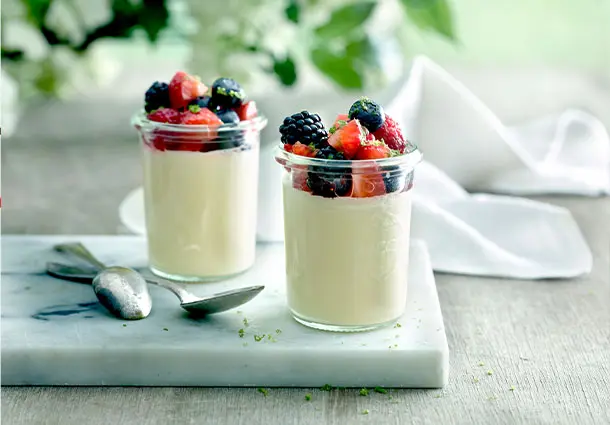 Gläser mit heller Panna cotta, darauf marinierte Beeren in rotem Sirup und feine Holunderblüten.