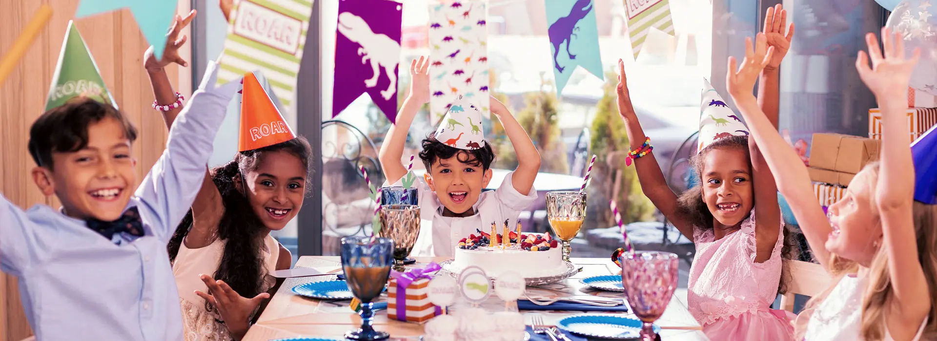Eine Gruppe fröhlicher Kinder sitzt an einem festlich gedeckten Tisch mit Geburtstagskuchen, Partyhüten und bunten Dekorationen.
