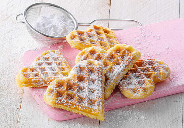 Herzförmige Waffeln liegen mit Puderzucker bestreut auf einem rosafarbenen Tablett
