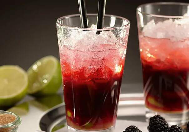 Auf einem grauen Tablett steht ein Glas, gefüllt mit einem roten Cocktails und Eiswürfeln. Daneben liegen Brombeeren und Limetten