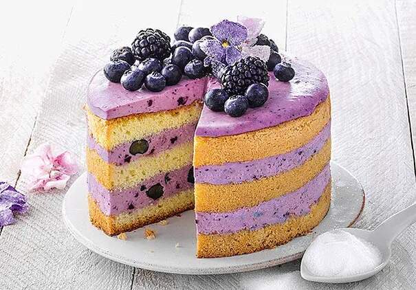 Eine hohe Heidelbeertorte zeigt abwechselnde Schichten aus goldgelbem Biskuitboden und violetter Beerencreme. Die Oberfläche ist mit frischen Heidelbeeren, Brombeeren und einer kandierten Veilchenblüte dekoriert; ein großes Stück ist herausgeschnitten, wodurch das Muster der Schichten sichtbar wird.