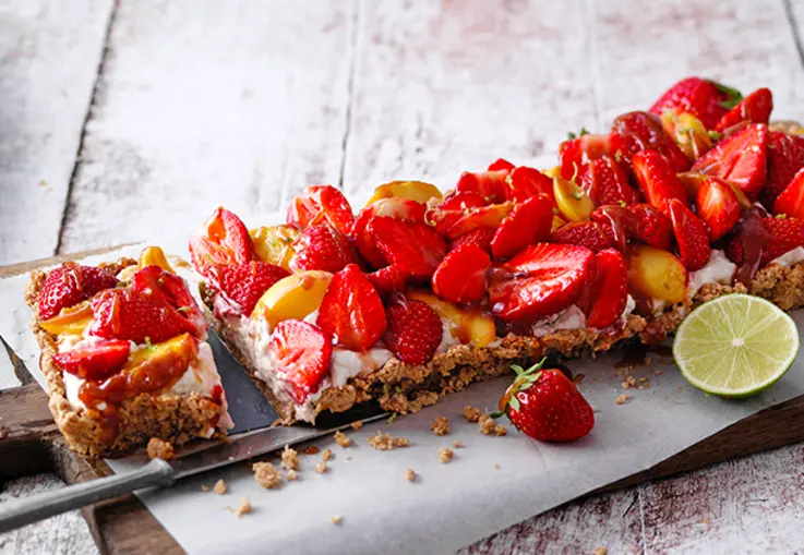 Eine Tarte mit nussigem Granola‑Boden und Limettencreme ist mit roten Erdbeeren, karamellisierten Pfirsichspalten und Nüssen garniert