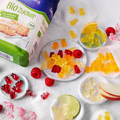 Auf einer weißen Tischdecke liegen viele bunte Gummibärchen verteilt, mit orangenscheiben und einer Tüte Zucker von Sweetfamily