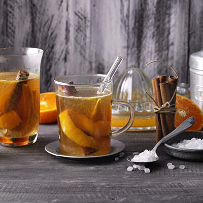 Drei Gläser mit heißem Tee sind mit Orangenscheiben, Zimtstangen und Sternanis aromatisiert; daneben stehen ein Teedose, eine Zuckerzange und ein kleiner Teller mit Zuckerkristallen.