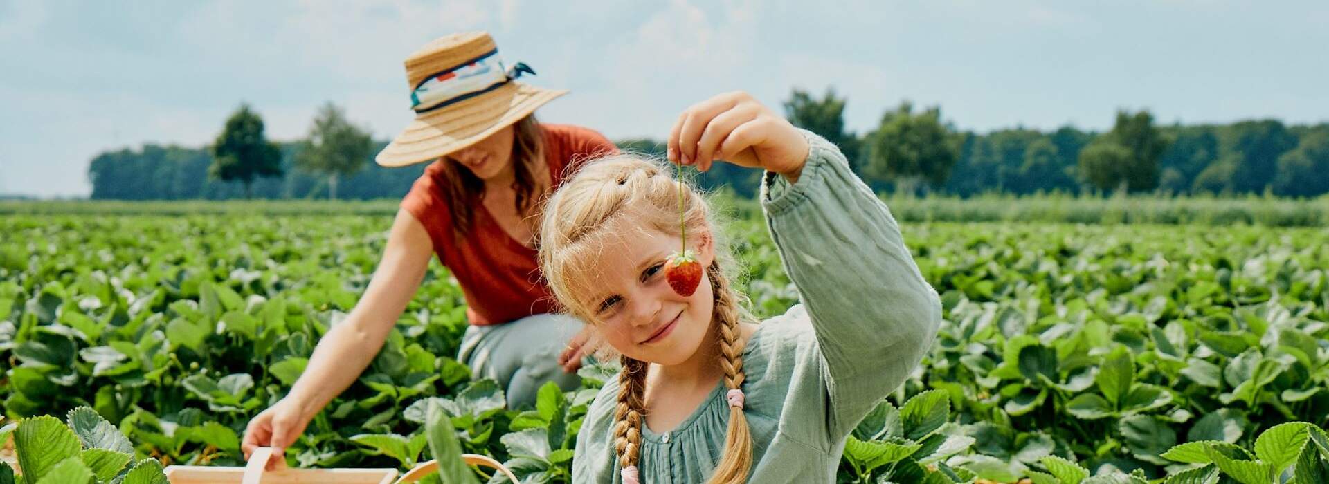 Ein Mädchen mit geflochtenem Haar pflückt eine Erdbeere auf einem Feld, eine Frau mit Hut ist im Hintergrund.