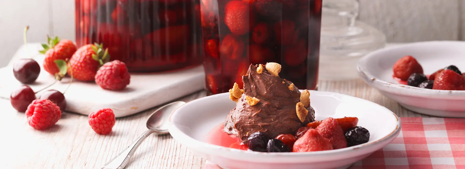 Im Vordergrund sthet ein kleiner Dessertteller mit einer Kugel Schokoeis darauf und mit Kekskrümeln und eingelegten Beeren.